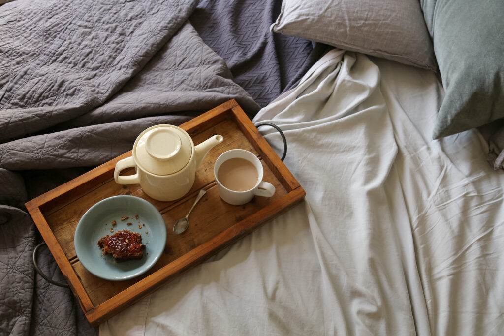 wooden breakfast tray on bed with teapot, tea and snack on a teal plate.  the bed features grey and cream linen sheets